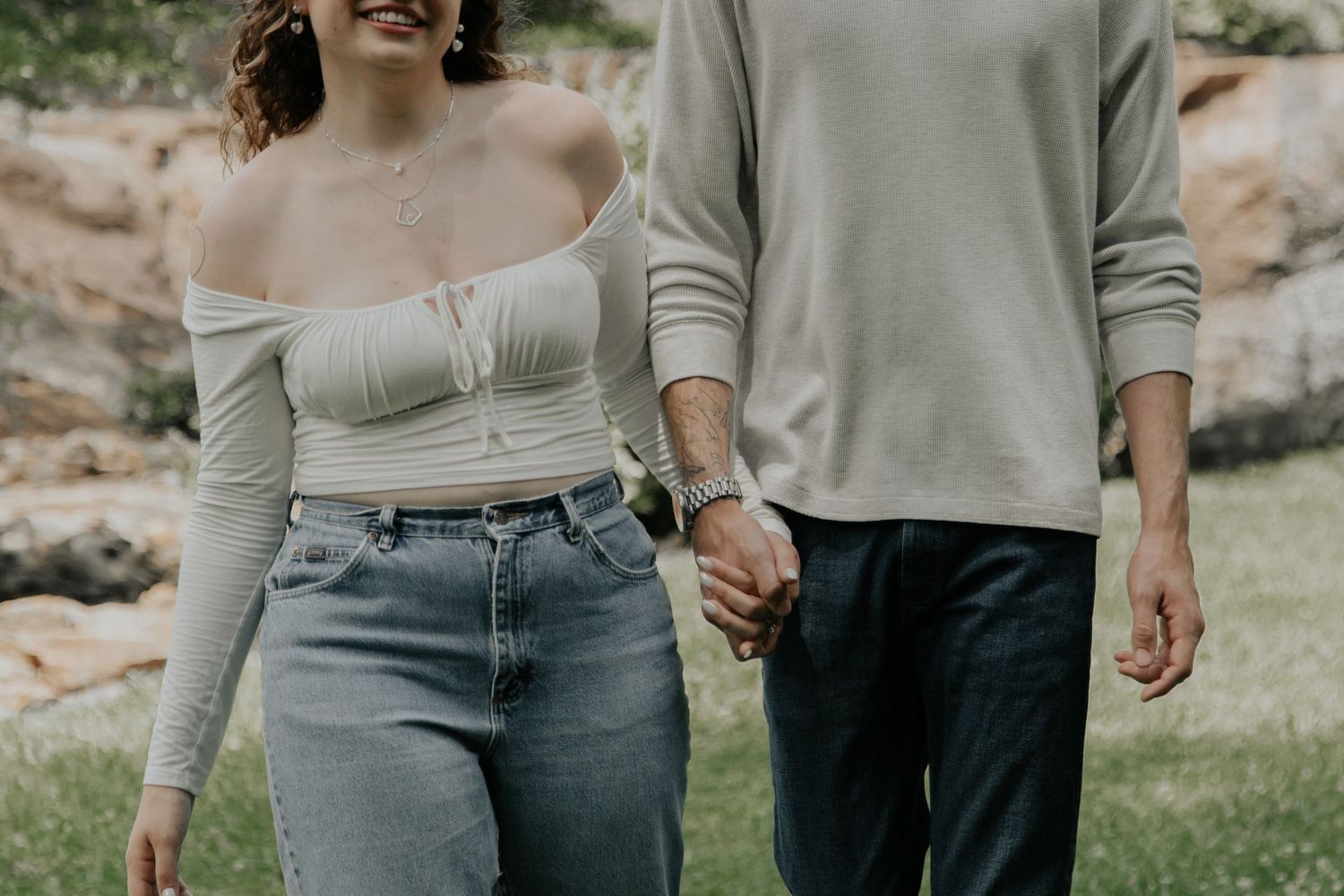 Couple holding hands while walking outdoors, woman in white crop top and jeans, man in gray sweater and dark pants.