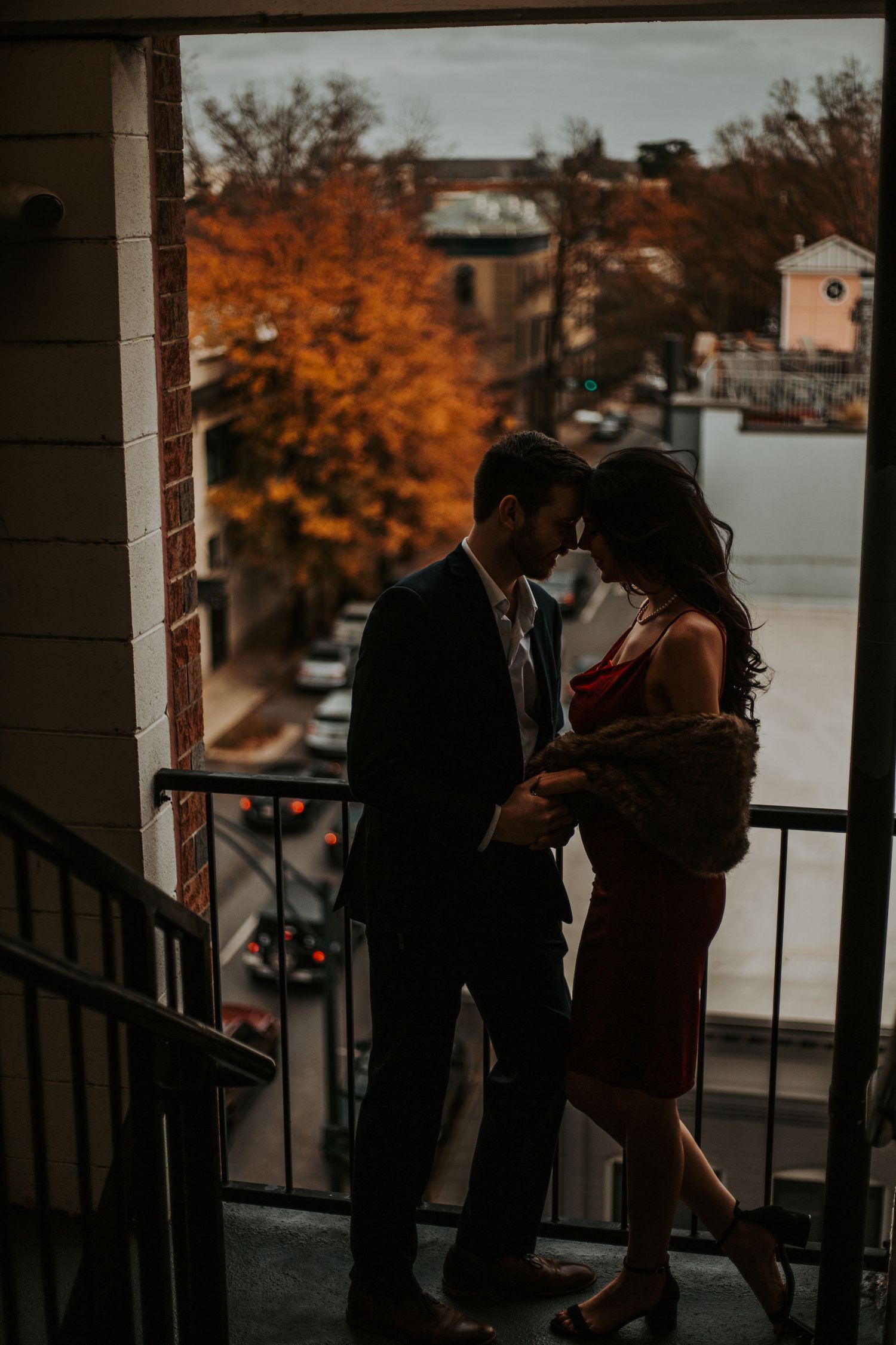Romantic couple embracing on a city balcony at dusk, surrounded by autumn foliage and urban street views.
