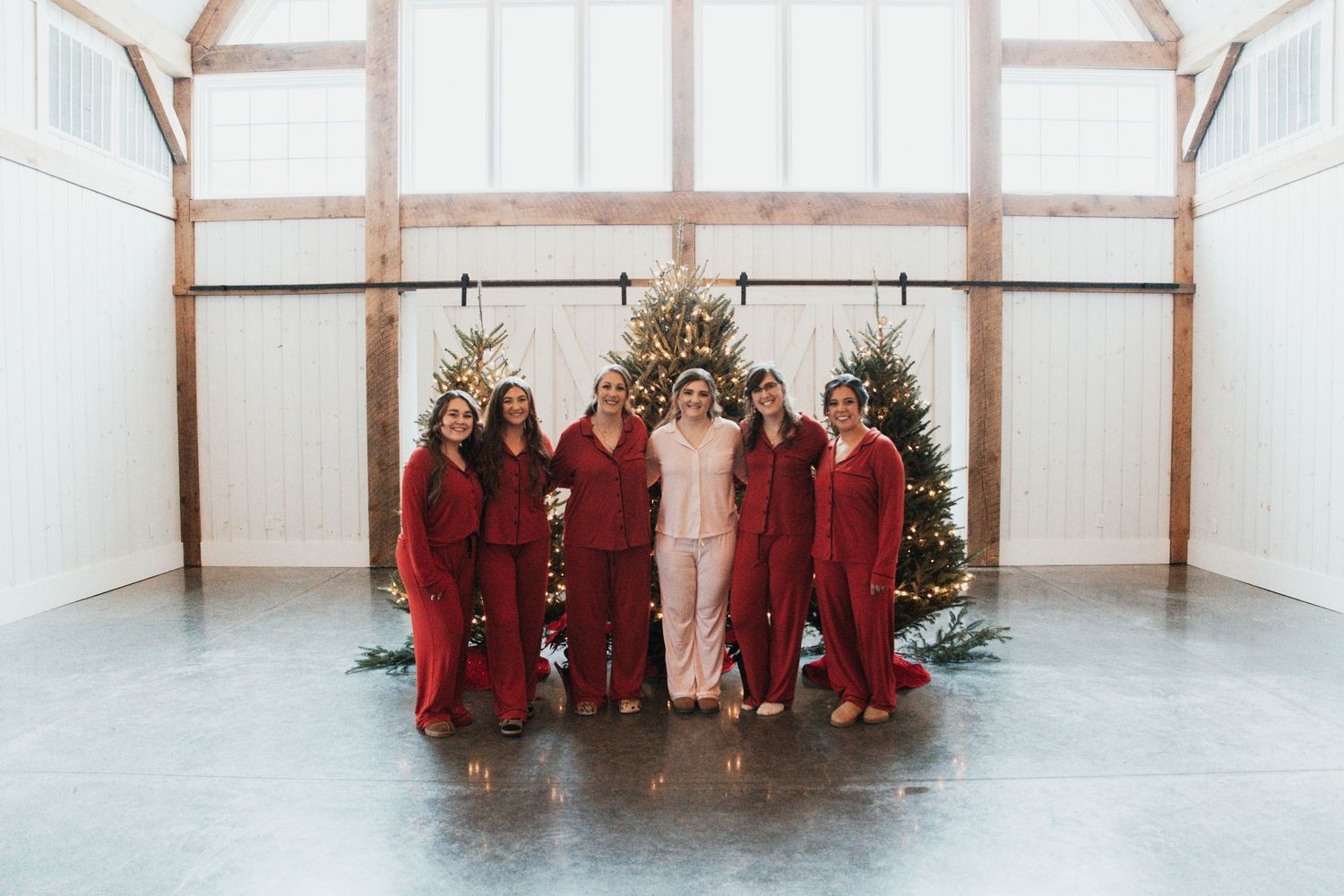 Bride in pink pajamas poses with bridesmaids in red jumpsuits inside a barn decorated with Christmas trees.