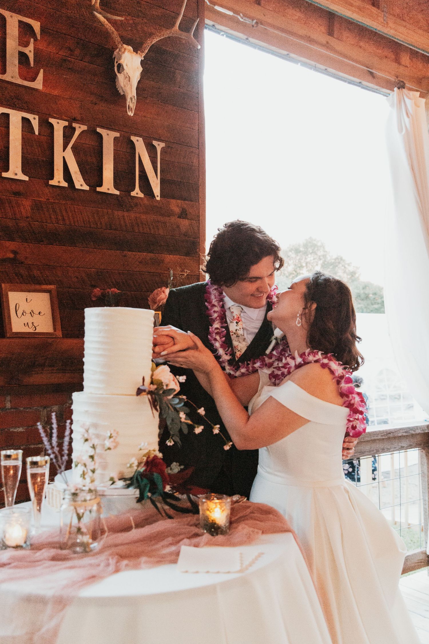 Bride and groom cutting white tiered wedding cake at rustic barn reception, wearing floral leis.