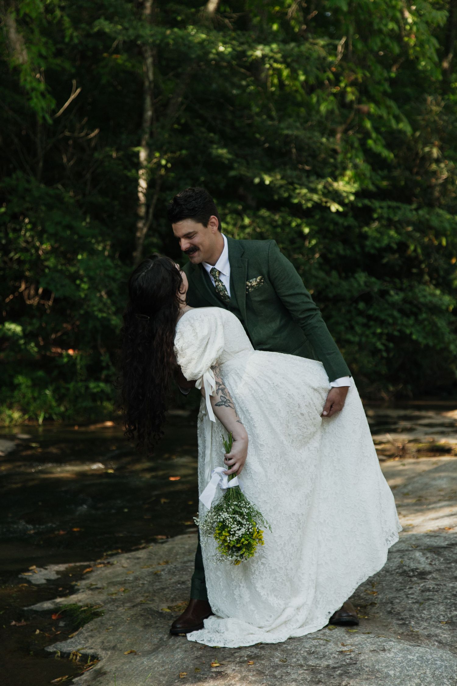 Groom in green suit dipping bride in white lace gown near sunlit forest creek, holding wildflowers. venue at ballenger bridge landrum sc