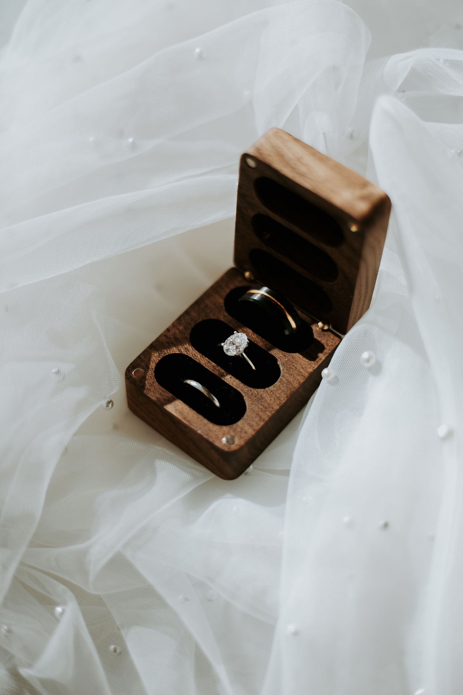 Elegant wooden ring box with wedding bands and diamond engagement ring displayed on white tulle bridal veil.