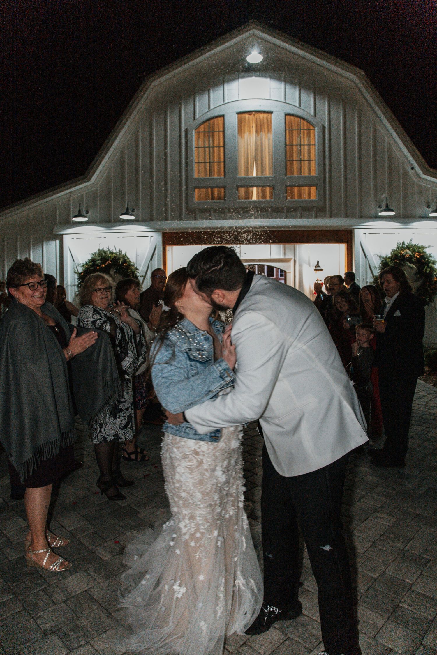 Newlyweds sharing a passionate kiss outside a white barn wedding venue surrounded by cheering guests at night. heyward manor (iva, sc)