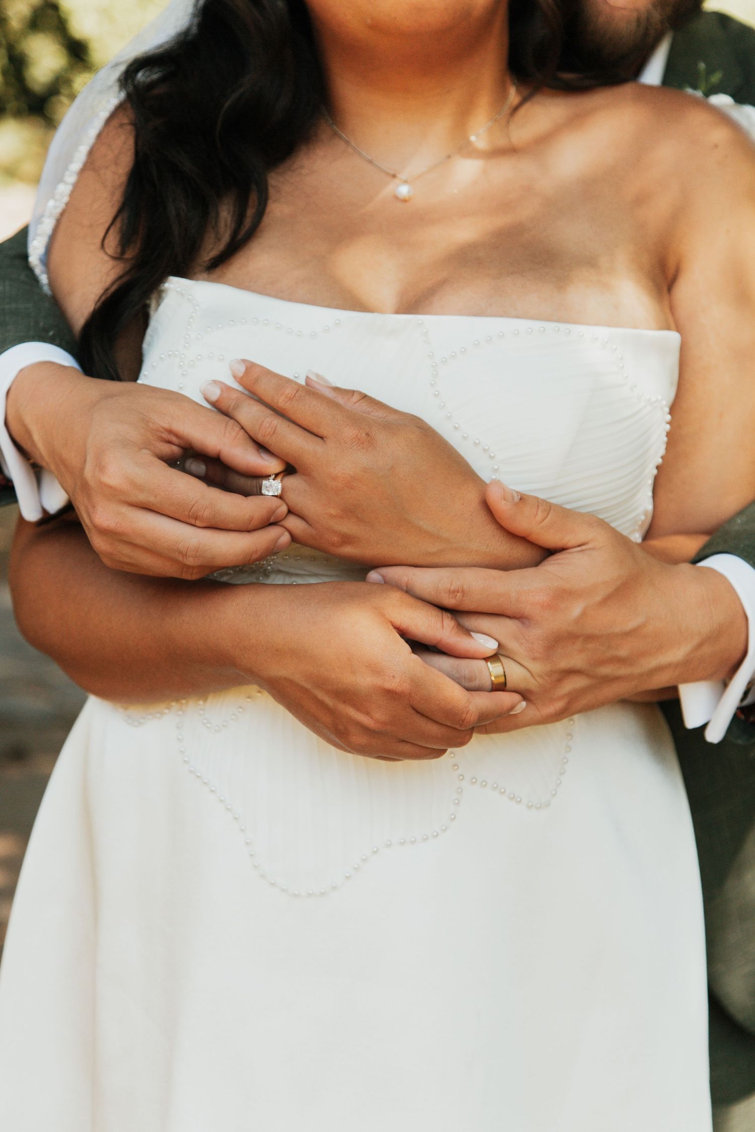 Close-up of bride and groom embracing, showcasing diamond engagement ring and gold wedding band on their hands.