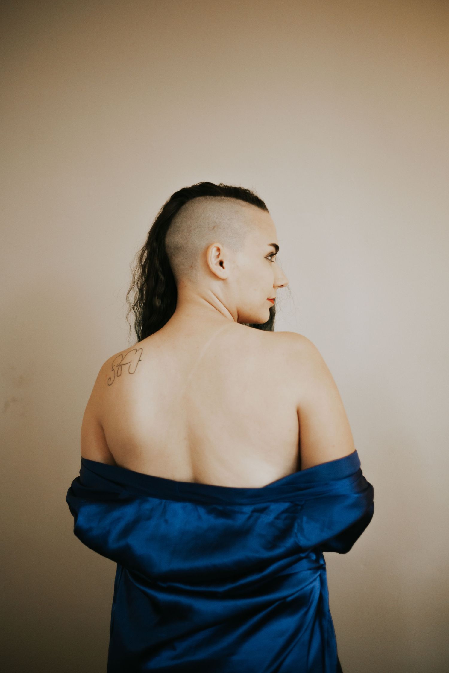 A person with a shaved undercut and tattoo on their back wears a blue satin off-shoulder garment.