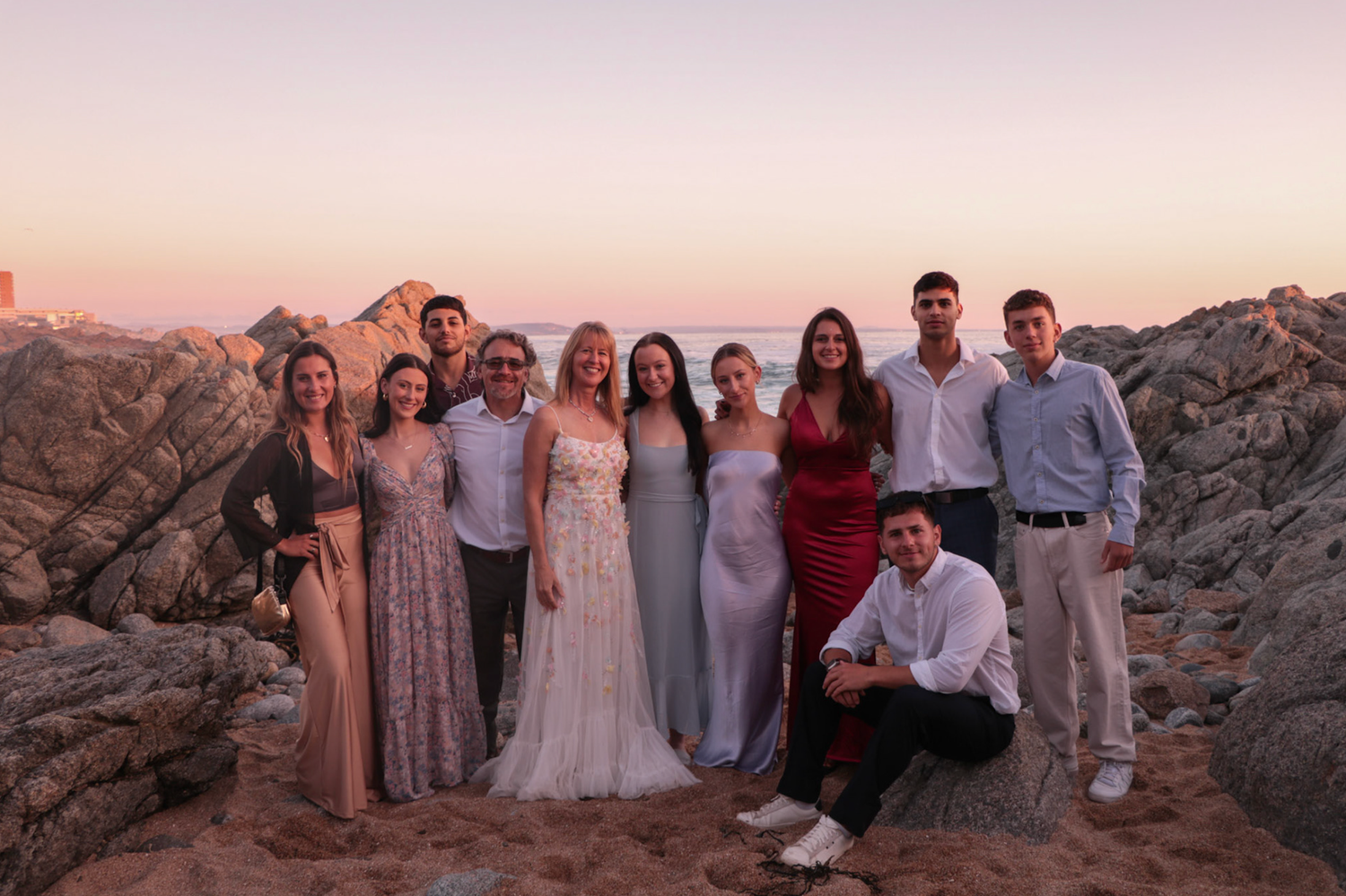 Wedding party group photo with bride, groom, and guests posing among rocks at golden hour sunset beach.