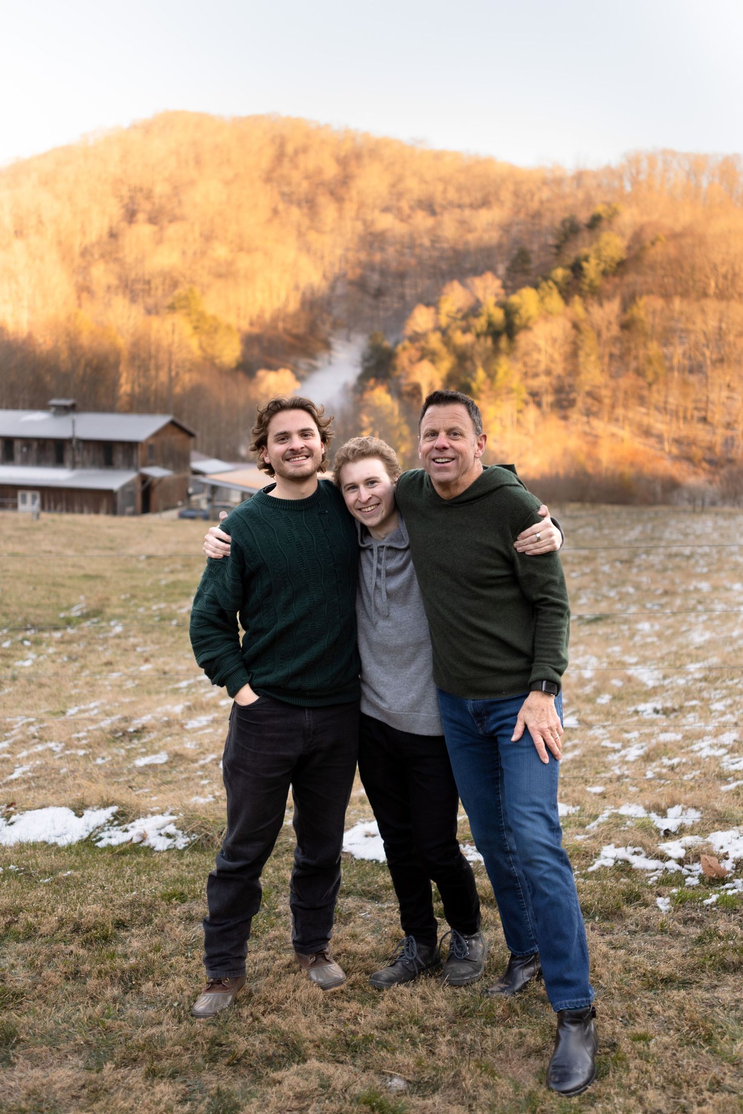 Three people smiling together outdoors in front of a mountain landscape with autumn foliage and light snow.