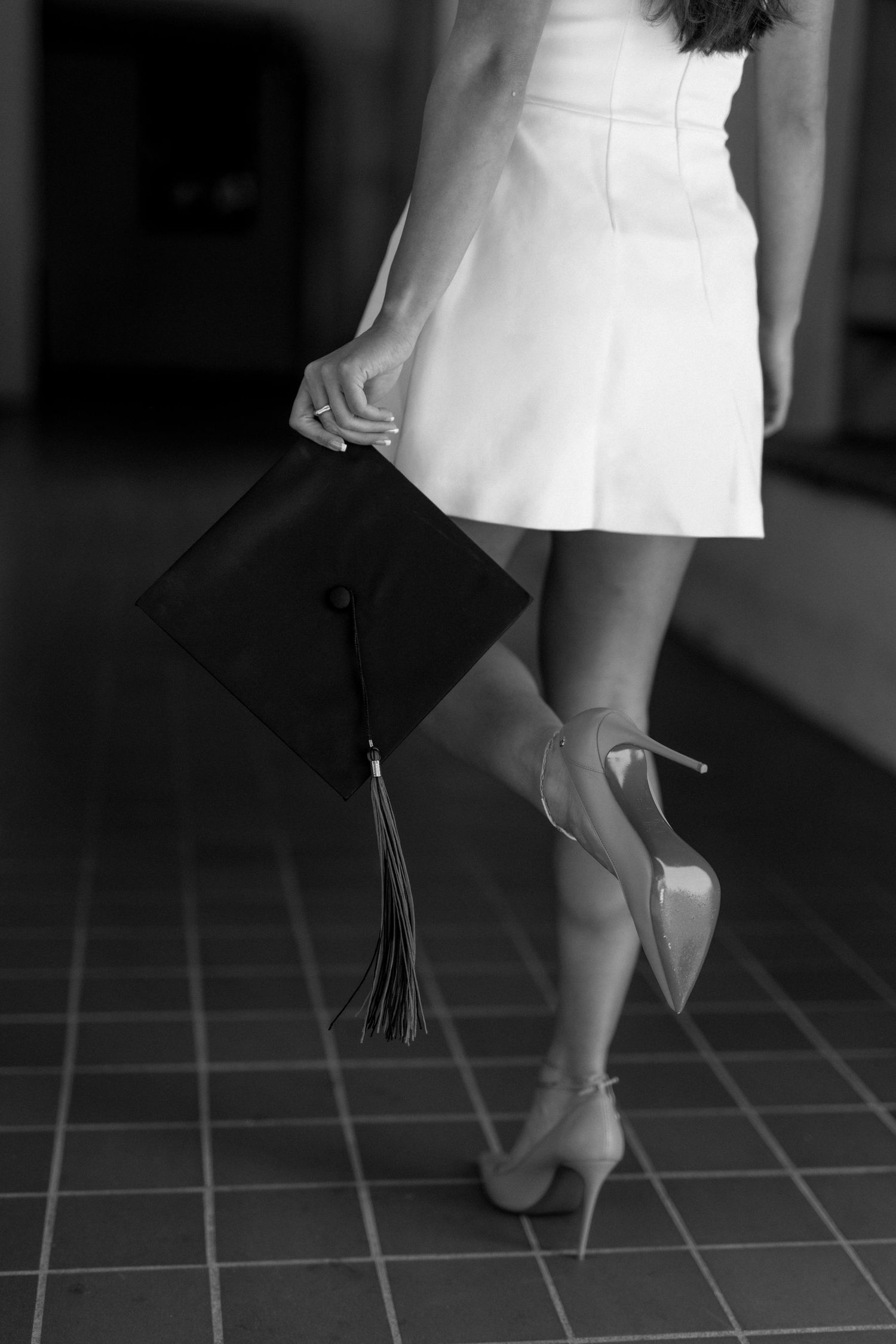 Woman in white dress holding graduation cap while wearing high heels, black and white photo.