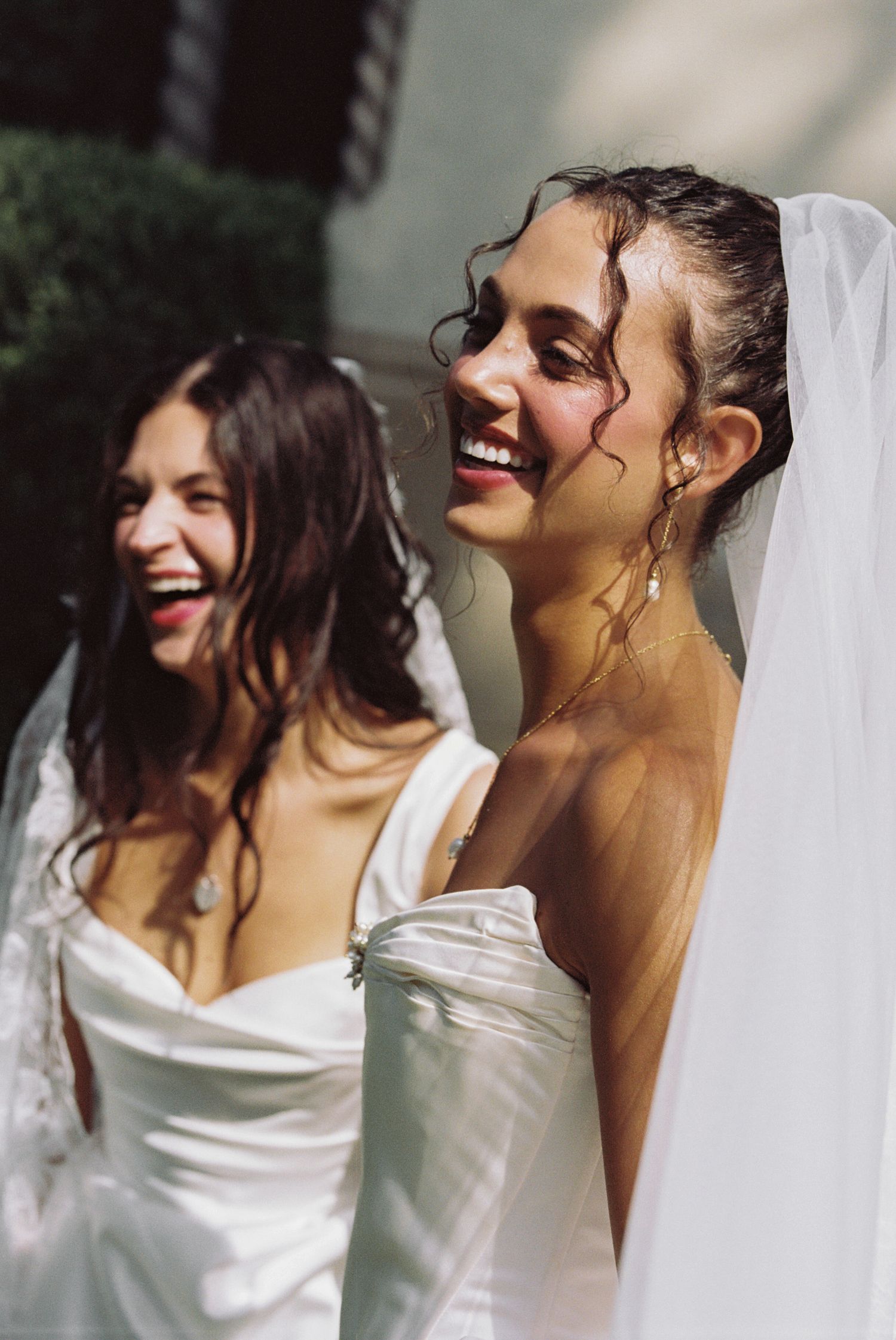 Two brides in white gowns and veils laughing joyfully outdoors on a sunny wedding day.