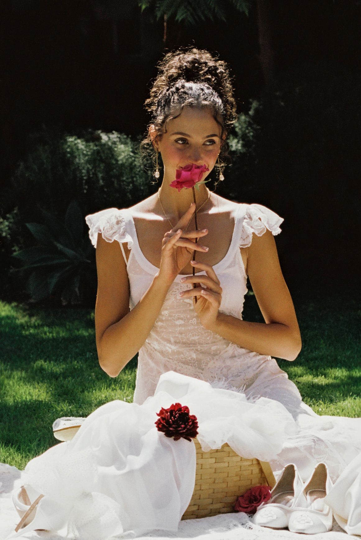 Bride in white dress sitting on grass, smelling a pink rose, with a wicker basket and white shoes nearby.