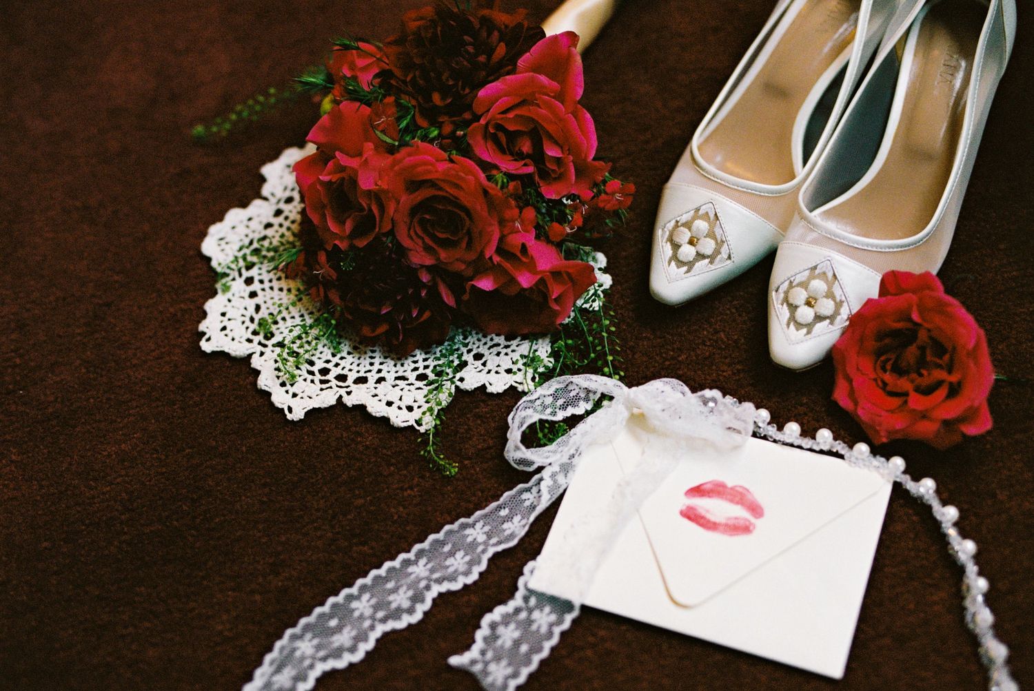 Bridal flat lay featuring red roses bouquet, white heels with jewels, lace garter, and envelope with lipstick kiss mark.
