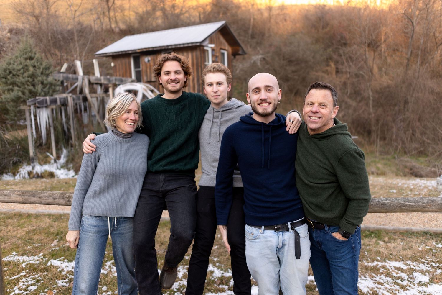 Five people smiling together outdoors in winter, standing in front of a rustic wooden barn with light snow on the ground.