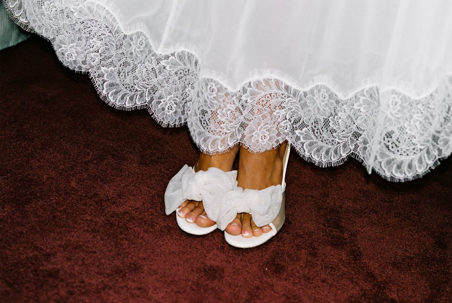 Close-up of bridal white heels with bow details beneath a lace wedding gown hem on red carpet.