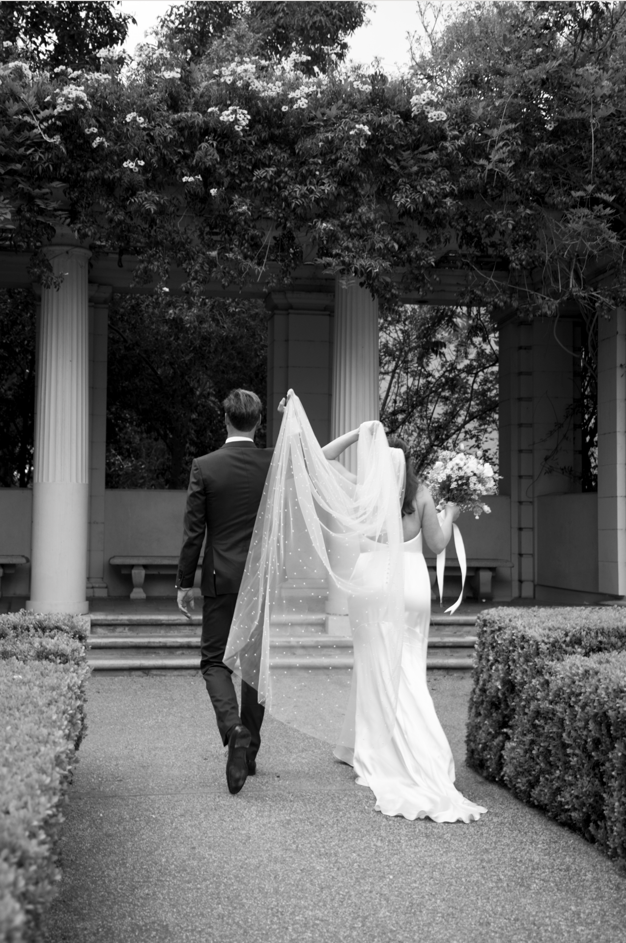 Black and white photo of bride and groom walking toward elegant columned garden venue, veil trailing behind.