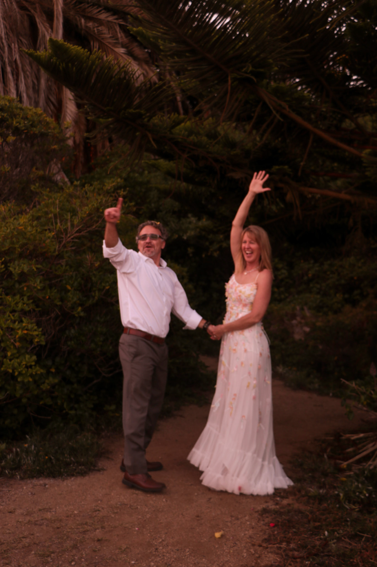Celebrating newlyweds hold hands and raise arms joyfully on garden path surrounded by lush tropical trees.