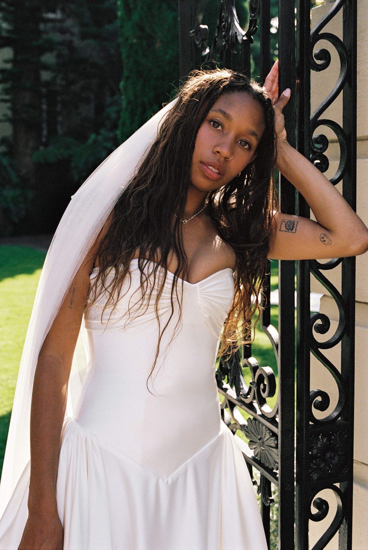 Bride in white strapless gown and veil poses by ornate black iron gate outdoors.