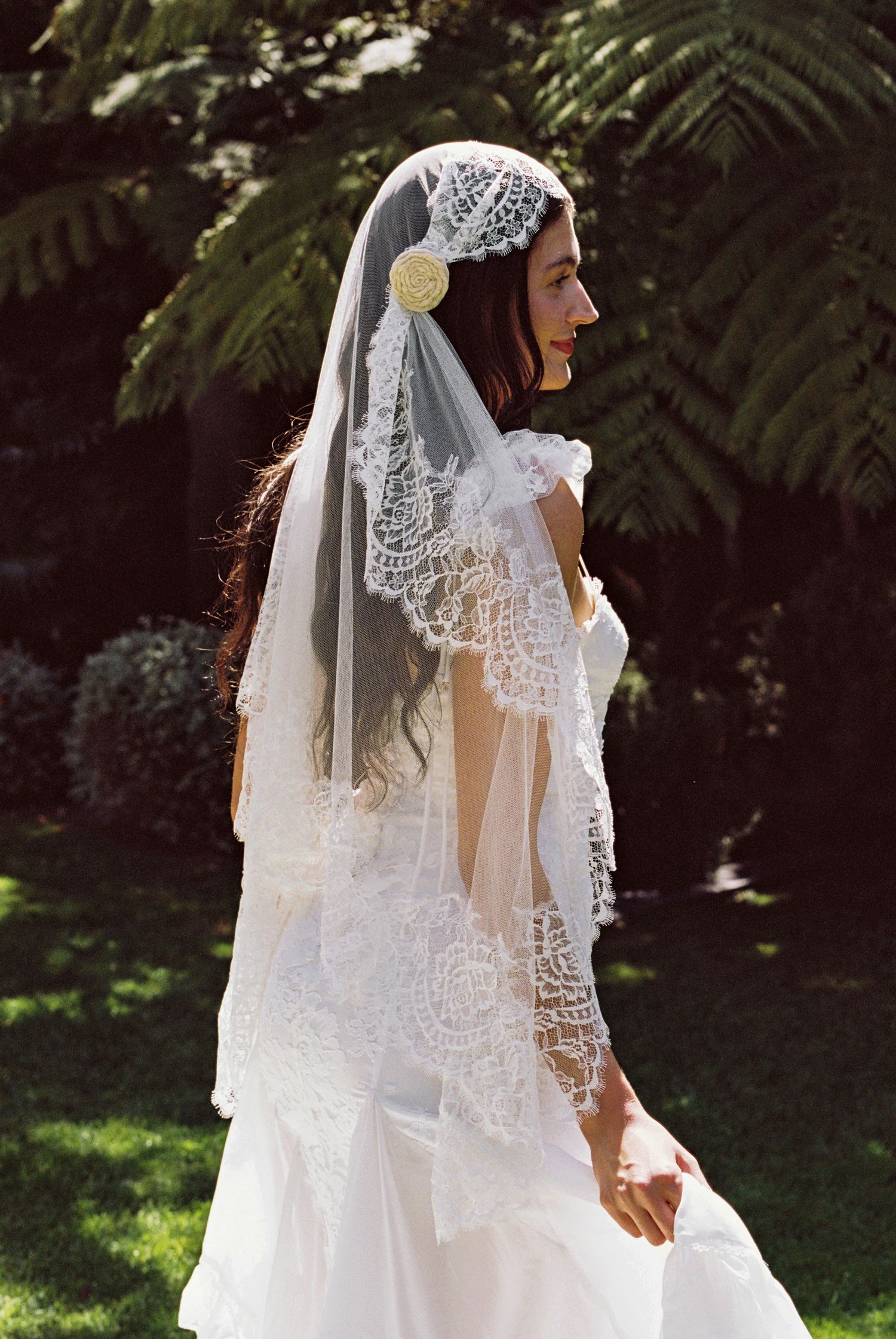 Bride wearing elegant lace veil with floral headpiece in garden setting, white wedding dress, lush green ferns background.