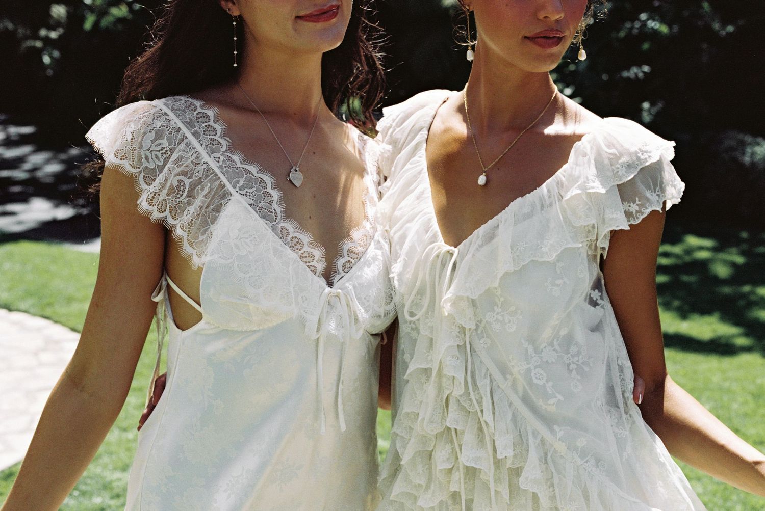 Two women wearing white lace ruffle dresses with V-necklines and cap sleeves, standing outdoors in natural light.