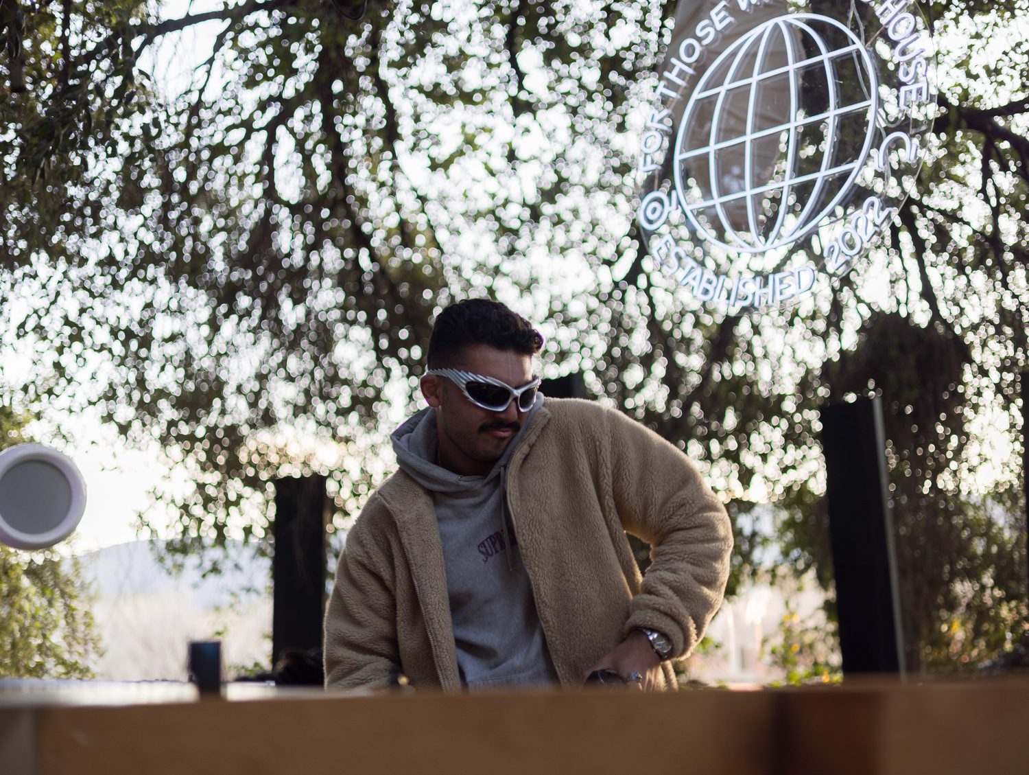 Male DJ in beige jacket and white sunglasses performing at outdoor daytime music festival.