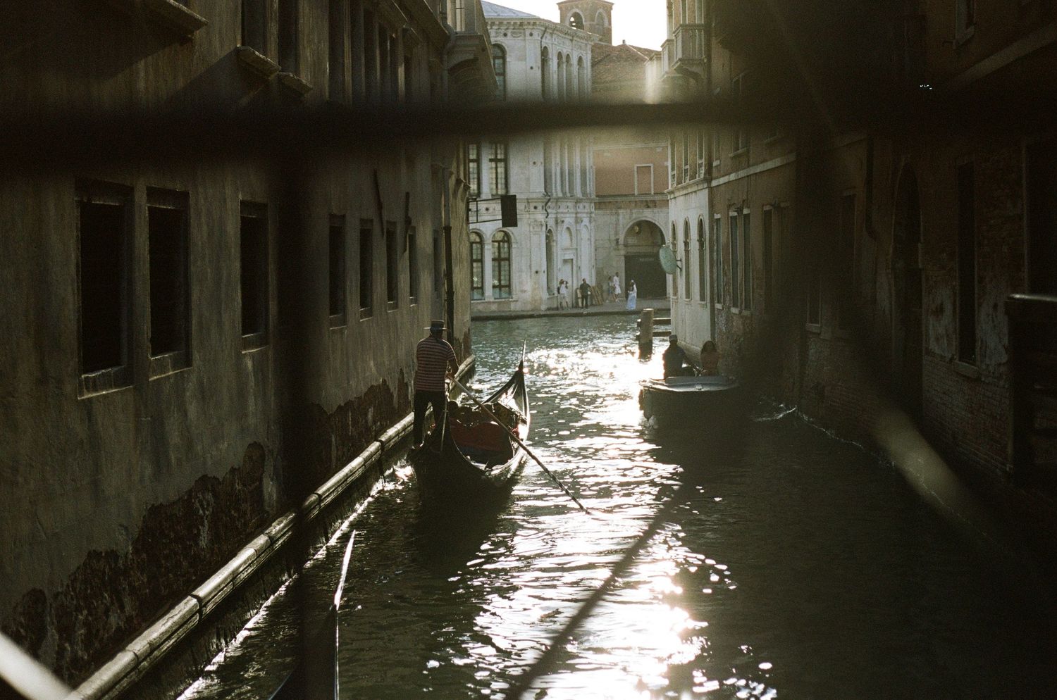 Gondolier navigating a narrow sunlit canal in Venice, Italy, with historic buildings reflecting on the water.