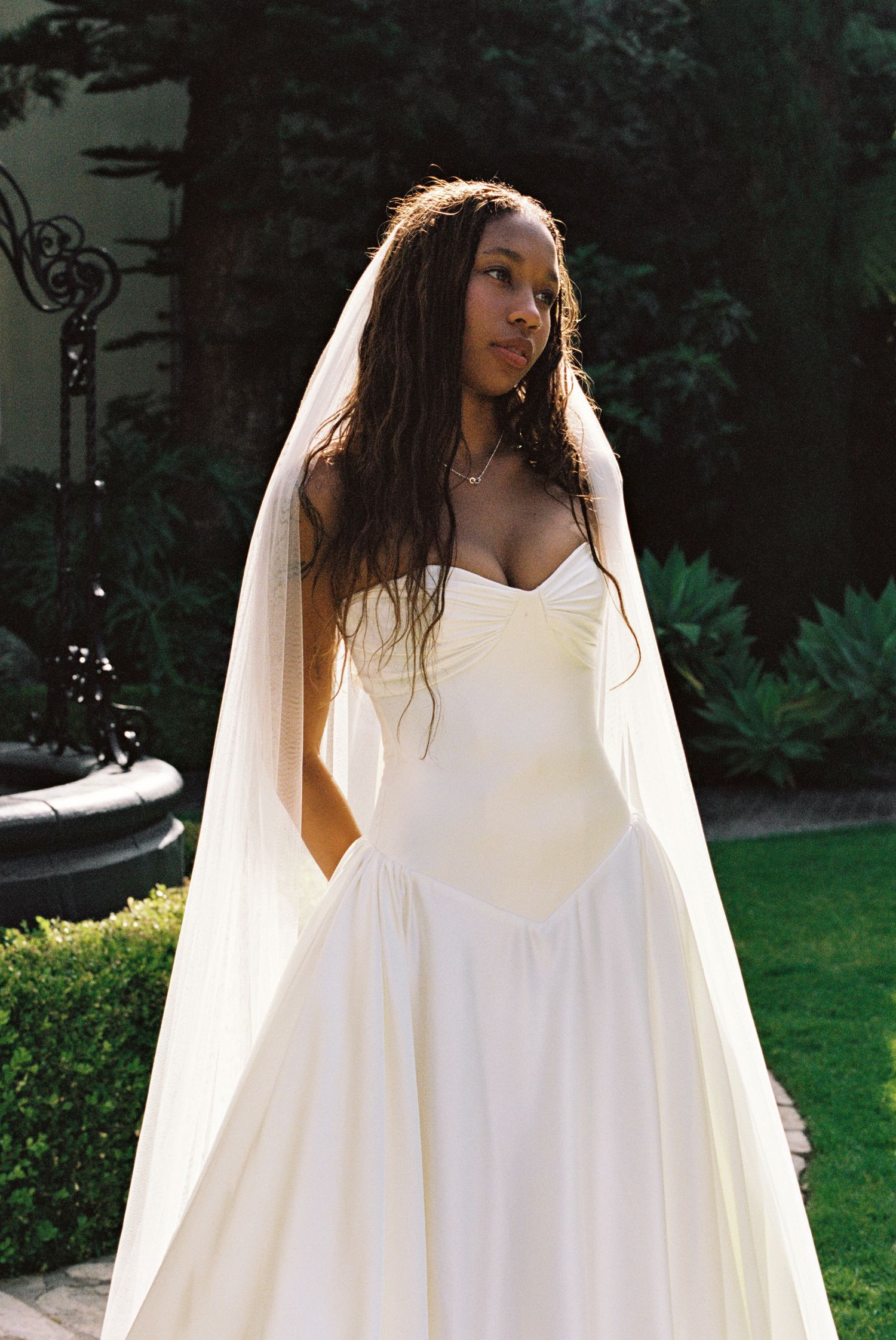 A Black bride in a strapless white gown and flowing veil stands outdoors in a lush garden setting.