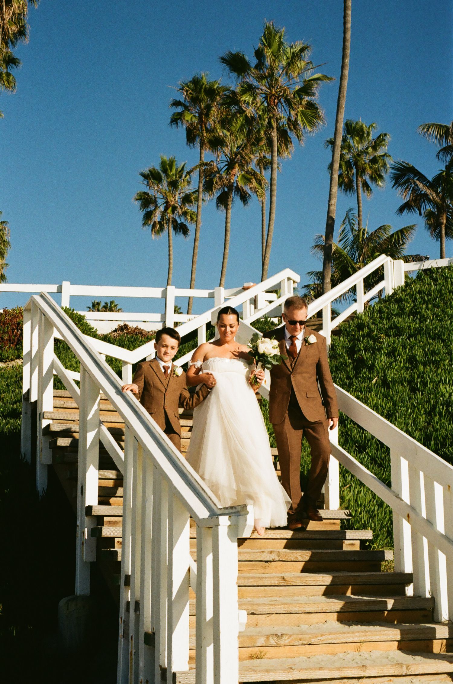 Bride walks down white wooden staircase flanked by groomsmen with palm trees in background.