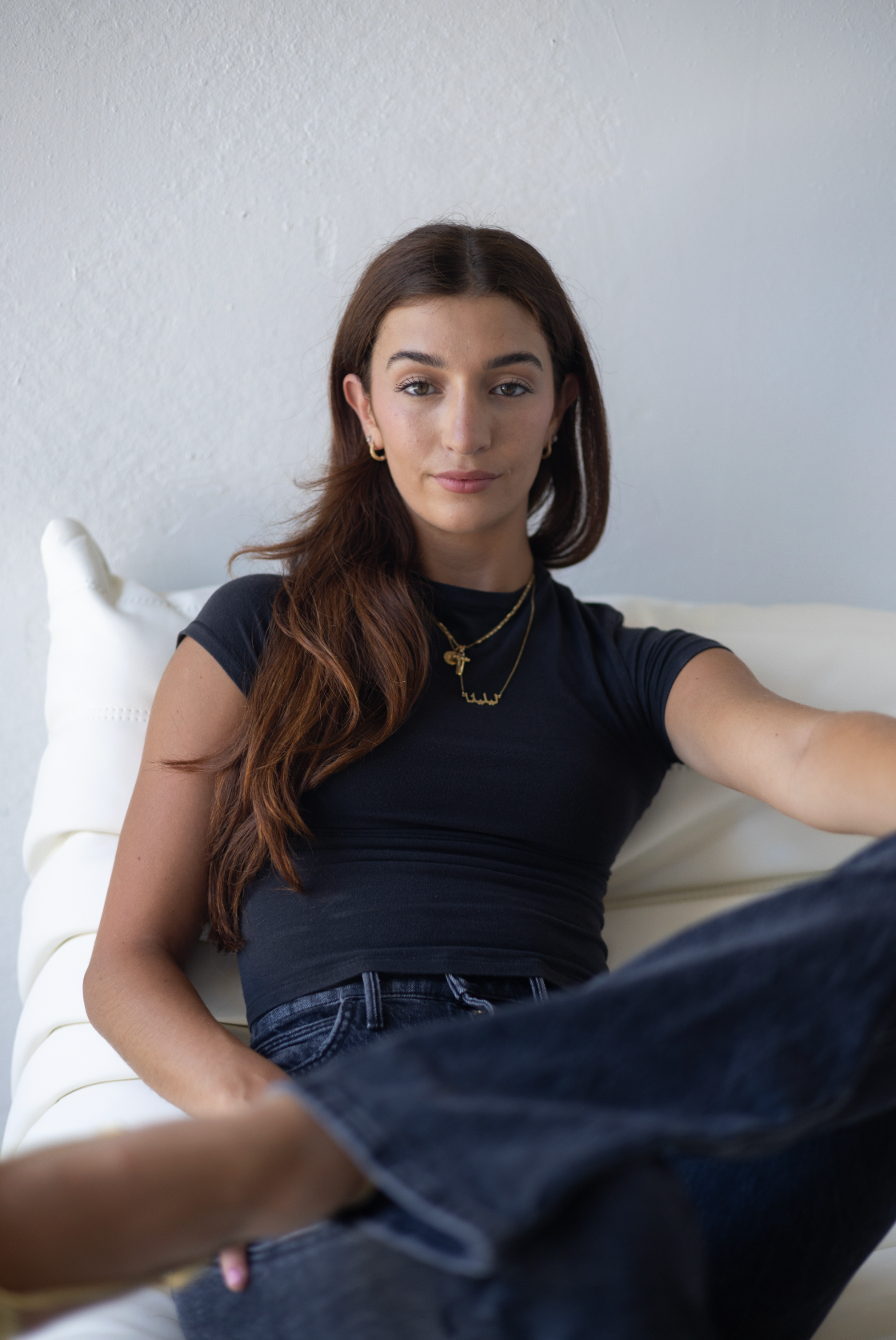 Young woman in black t-shirt and jeans wearing gold necklaces, relaxing on white chair.