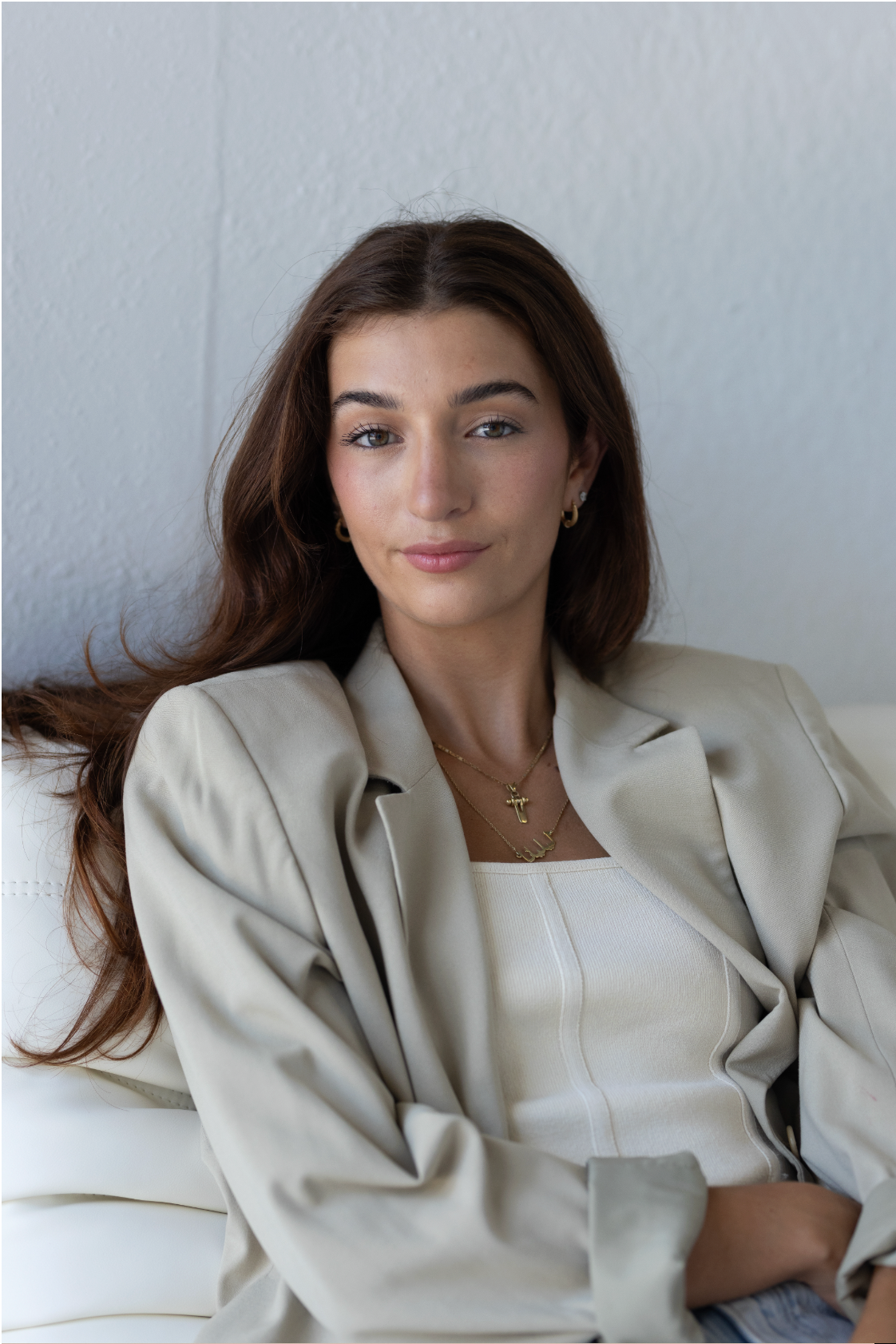 Young brunette woman in beige blazer relaxing on white sofa with gold necklace indoors.