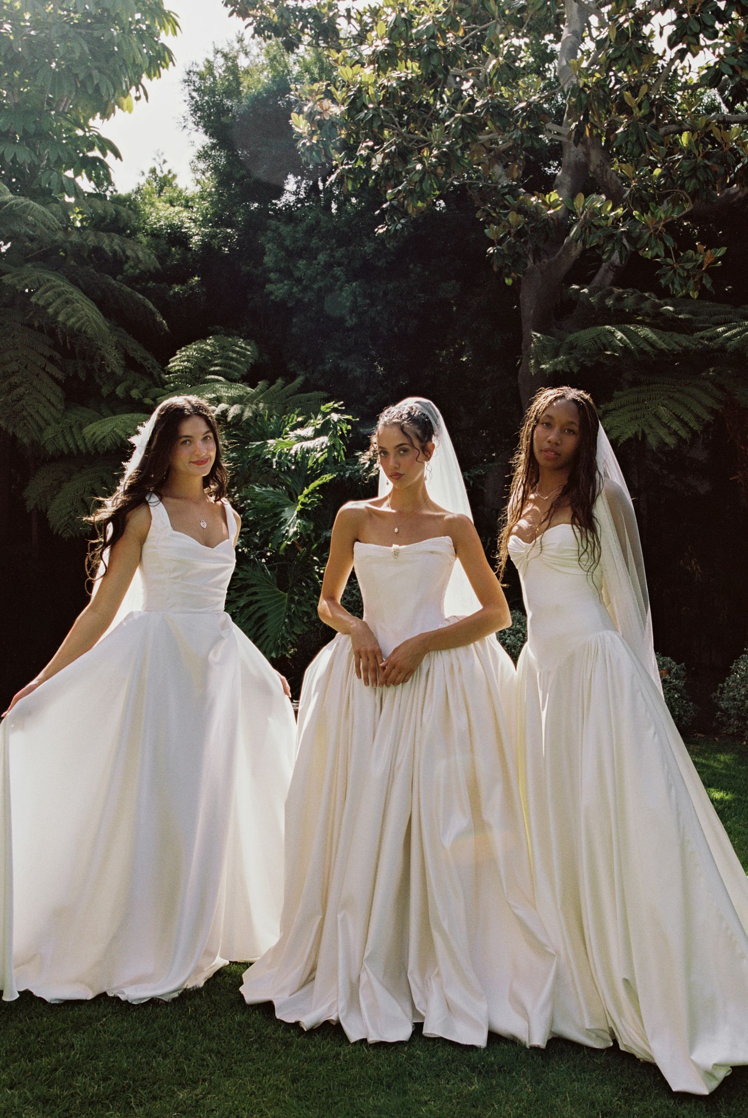 Three women in elegant white bridal gowns and veils pose outdoors in a lush garden setting.
