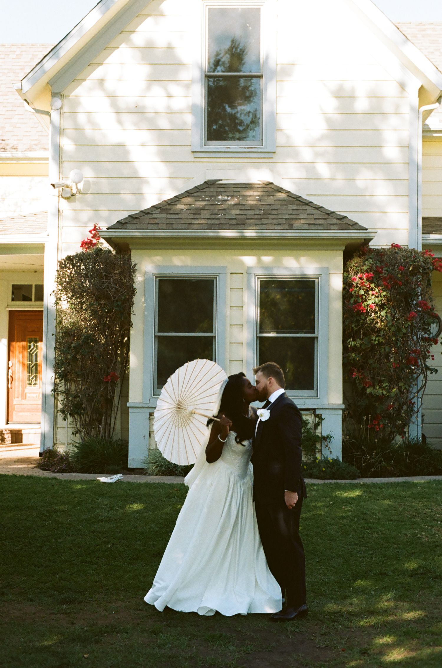 Bride and groom kissing in front of white farmhouse, bride holding decorative parasol umbrella.