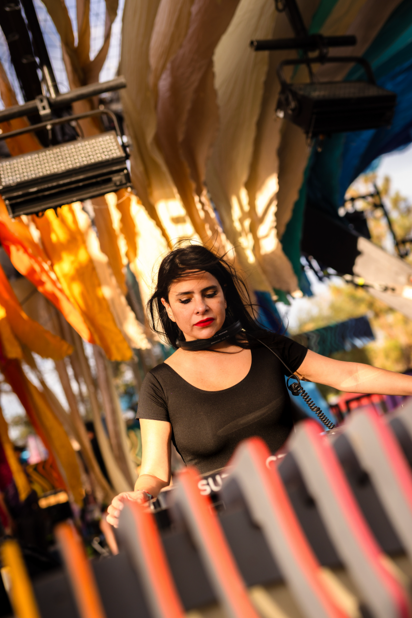 Female DJ in black top performing at an outdoor festival stage with colorful fabric backdrop.