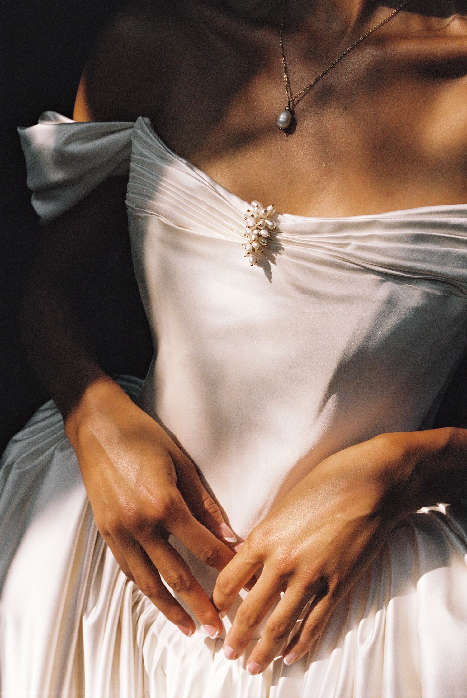 Bride in white off-shoulder gown with floral brooch and pearl necklace, hands clasped at waist.