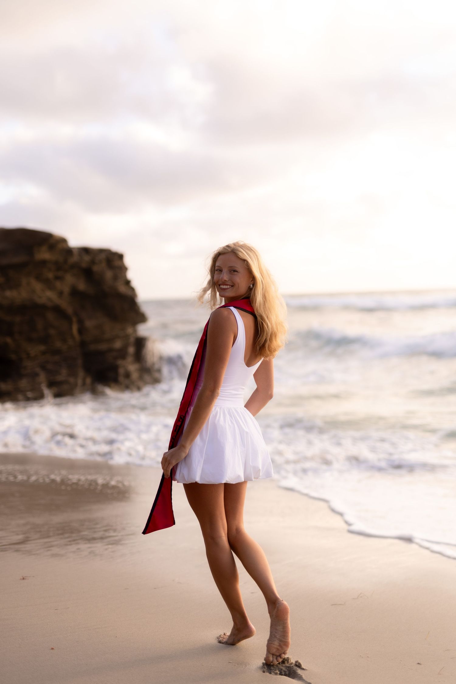 Smiling blonde woman in white dress holding red accessory on sandy beach with ocean waves and rocky cliffs.