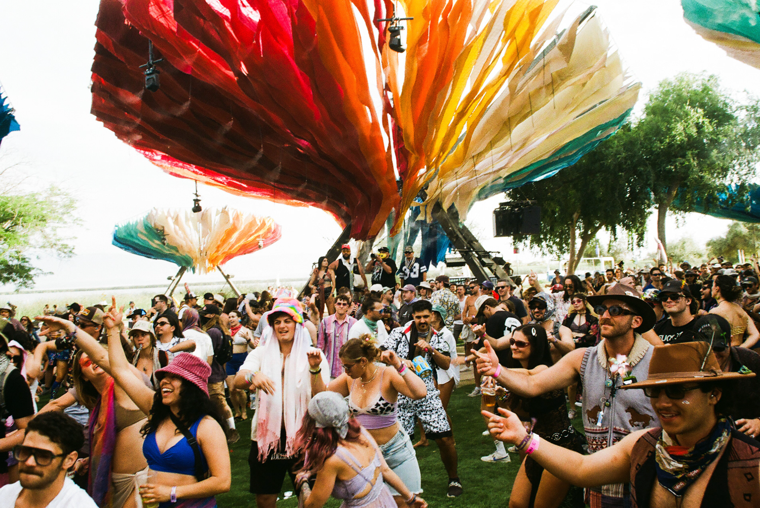 Colorful outdoor music festival crowd under vibrant hanging fabric decorations on a sunny day.