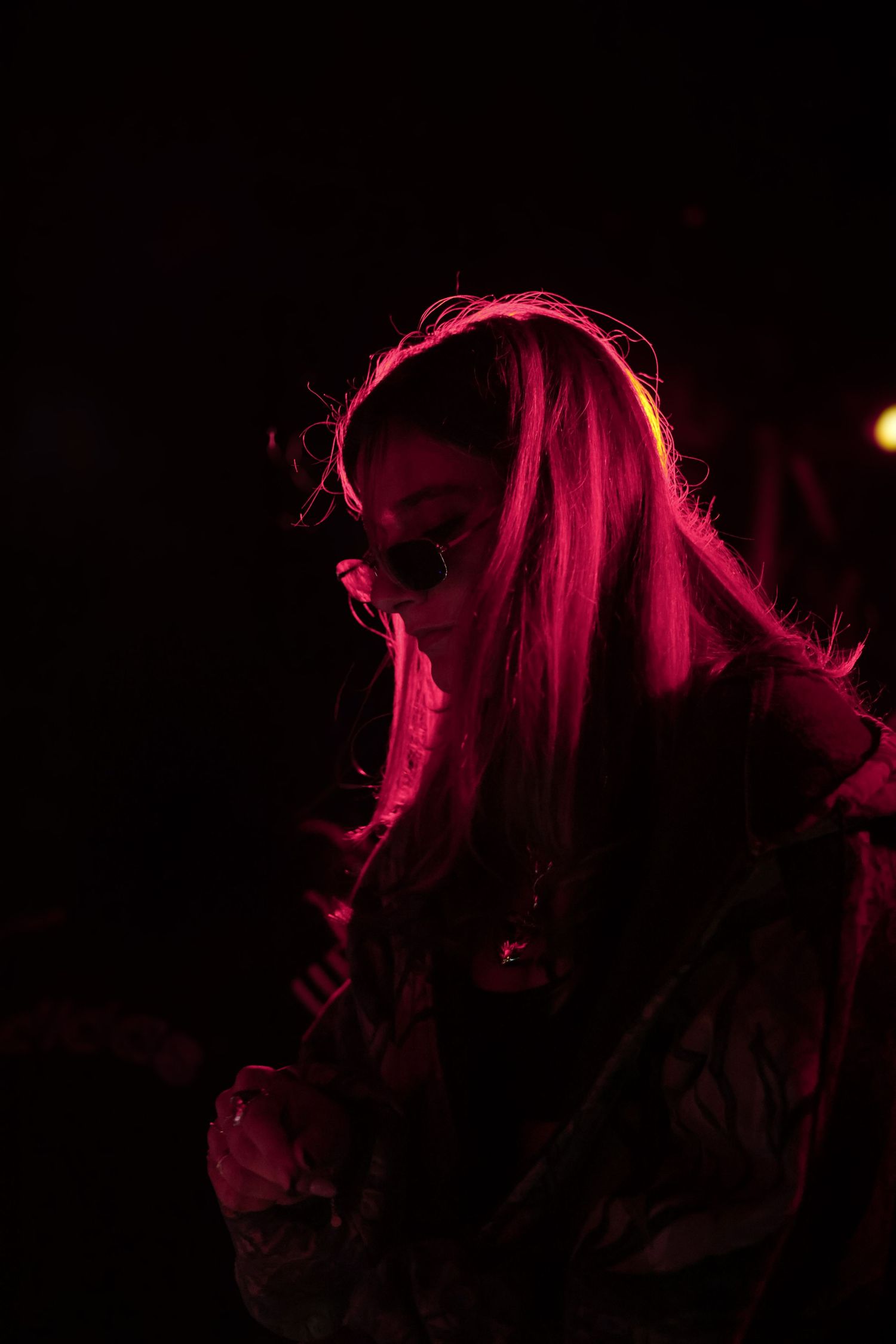 Woman with long hair lit by red neon light wearing sunglasses at dark concert.
