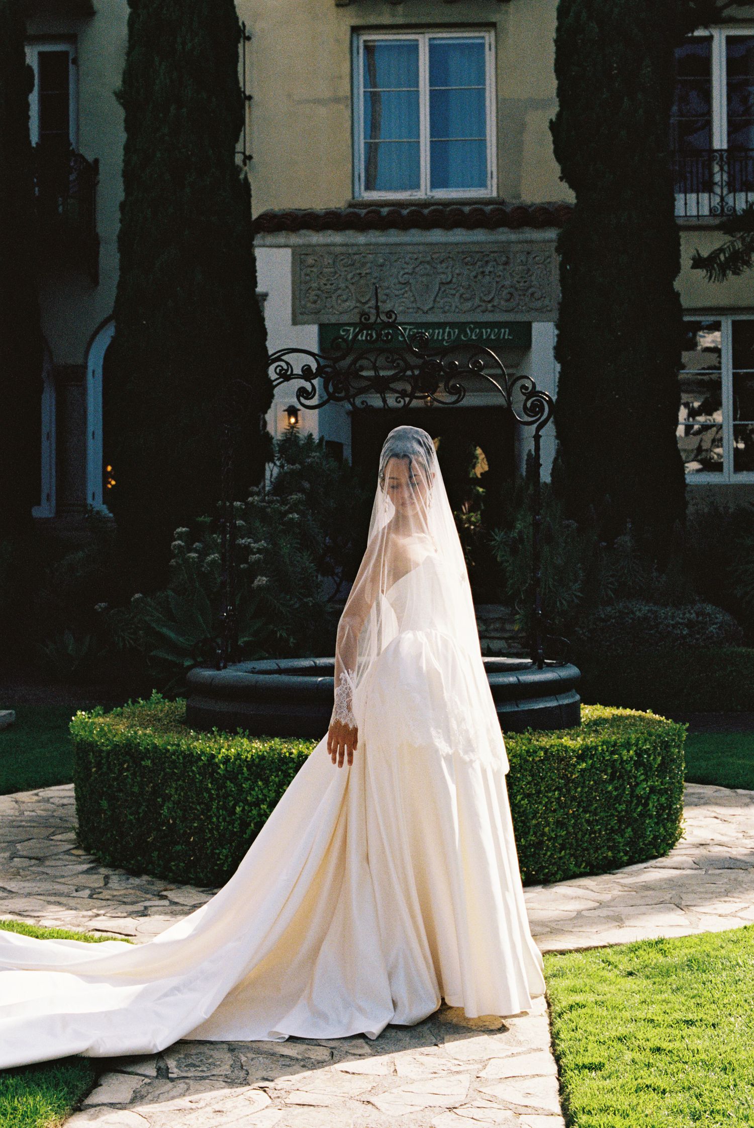 Bride in elegant white gown with long cathedral veil stands on garden path before a grand estate building.