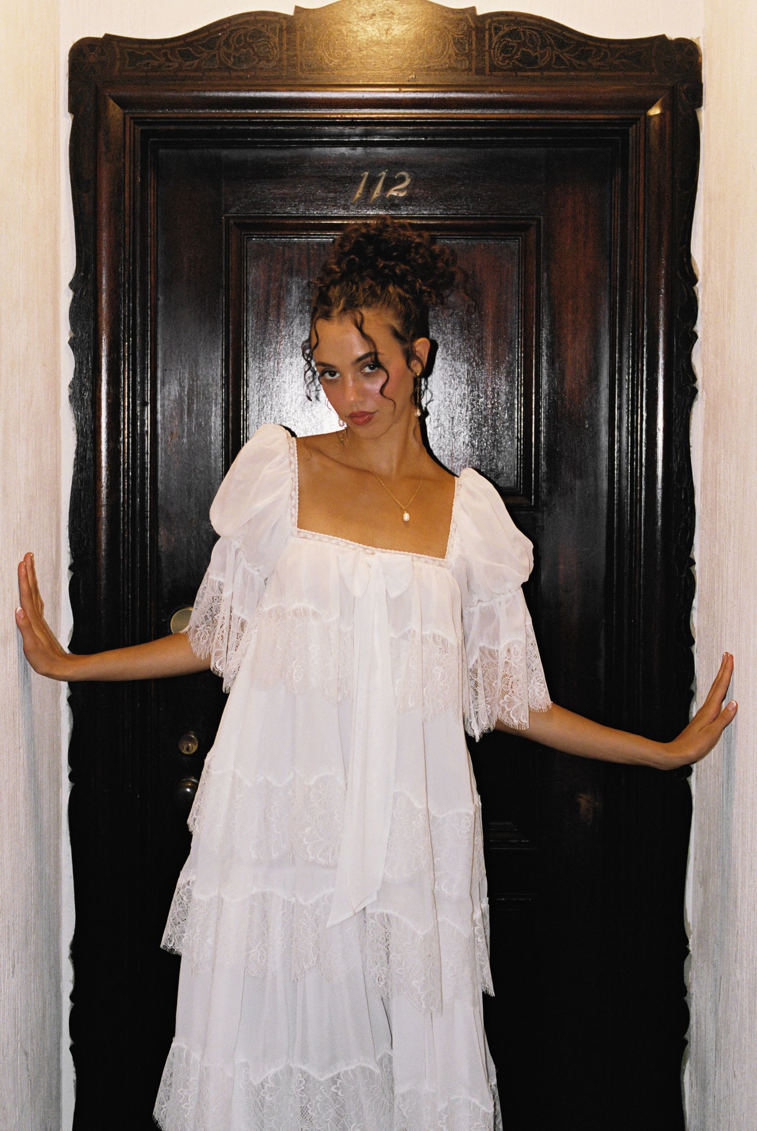 Woman in white lace tiered dress posing in front of dark wooden door numbered 112.