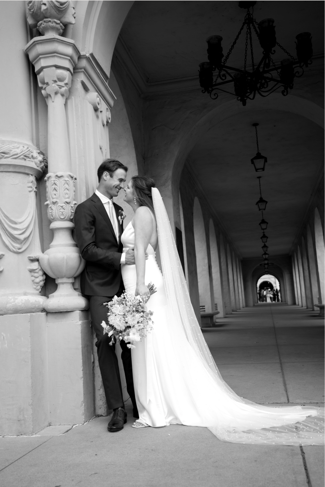 Black and white romantic photo of newlyweds sharing a kiss under ornate arched colonnade at Balboa Park.