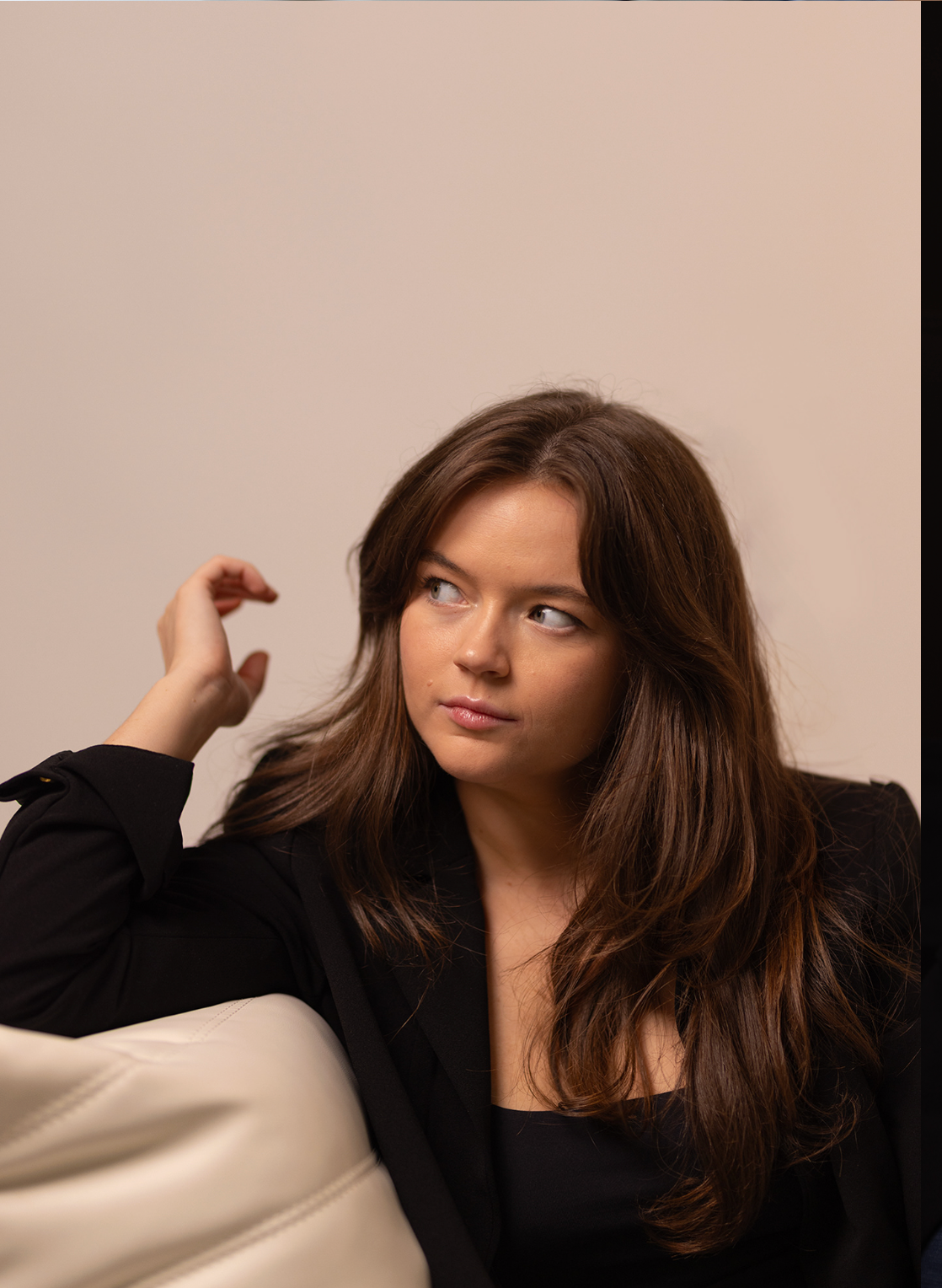 Brunette woman in black blazer looking away thoughtfully while seated on cream sofa.