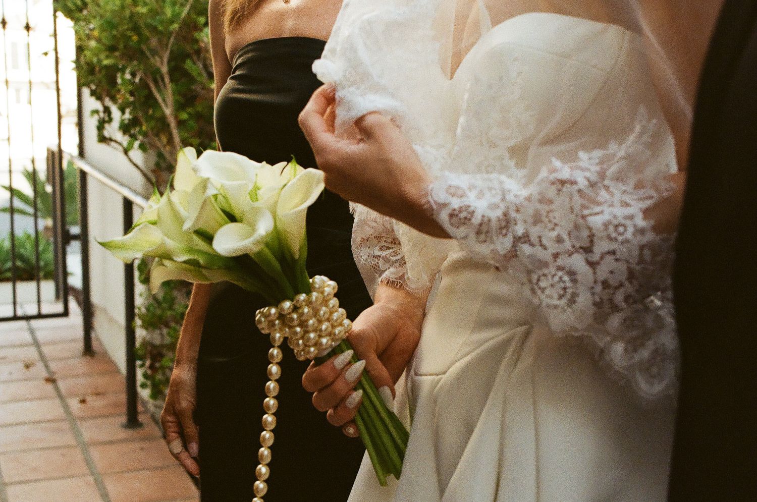 Bride in lace wedding gown holding white calla lily bouquet wrapped with pearl bracelet beside bridesmaid.