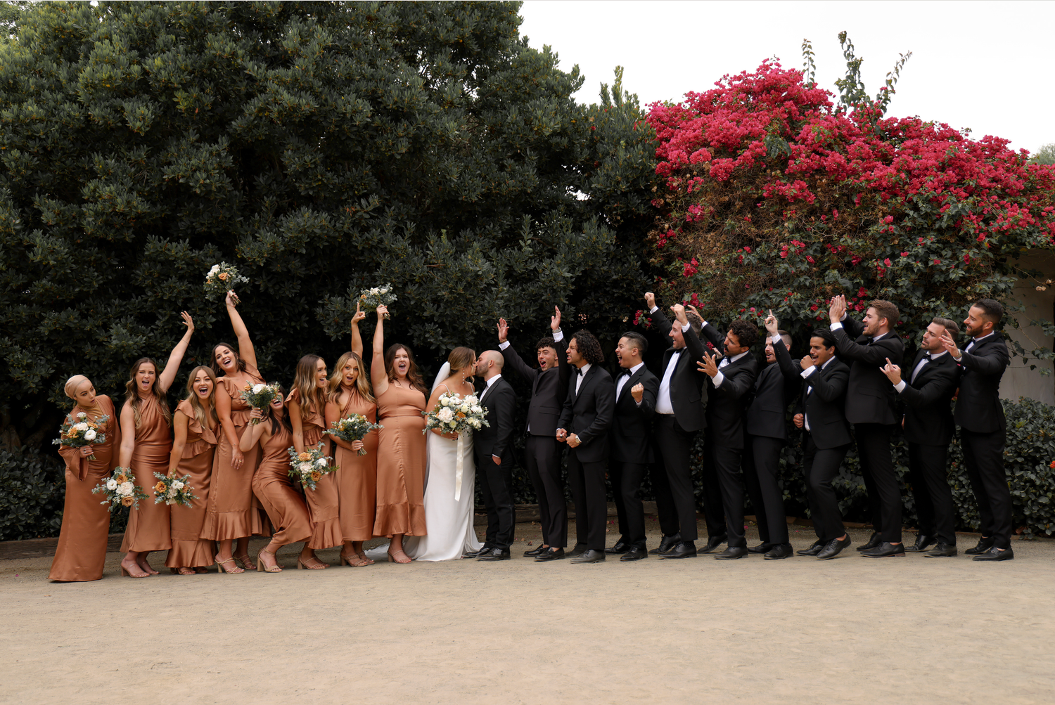 Wedding party celebrating outdoors, bridesmaids in rust satin dresses and groomsmen in black suits near blooming red flowers.