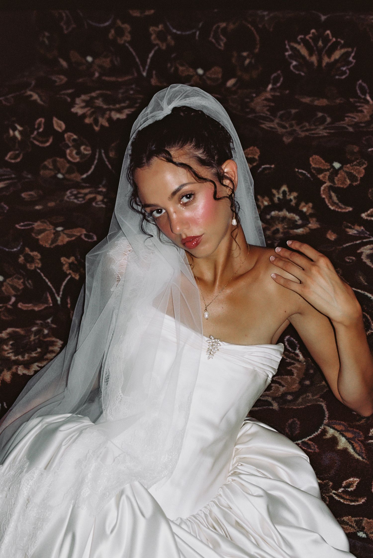A young bride in a white strapless gown and veil poses against a dark floral backdrop with elegant jewelry.