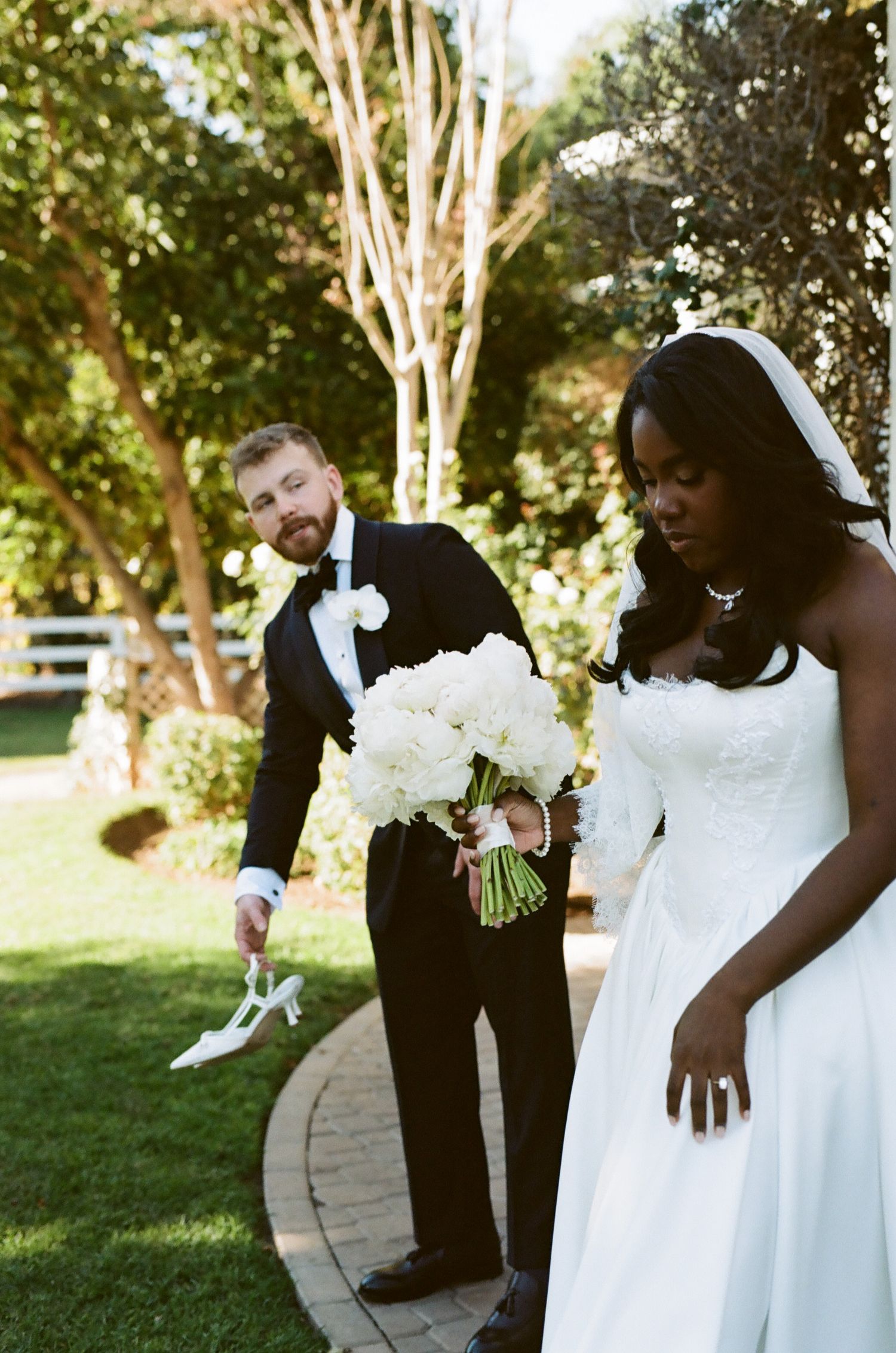 Groom in black tuxedo carries bride's heels while bride holds white peony bouquet outdoors.