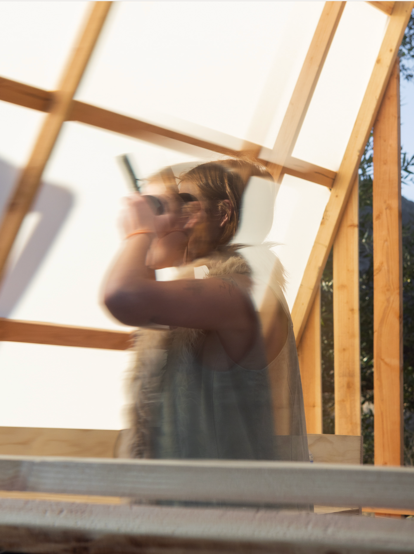 Blurred artistic wedding photo of woman drinking through wooden frame structure in warm sunlight.