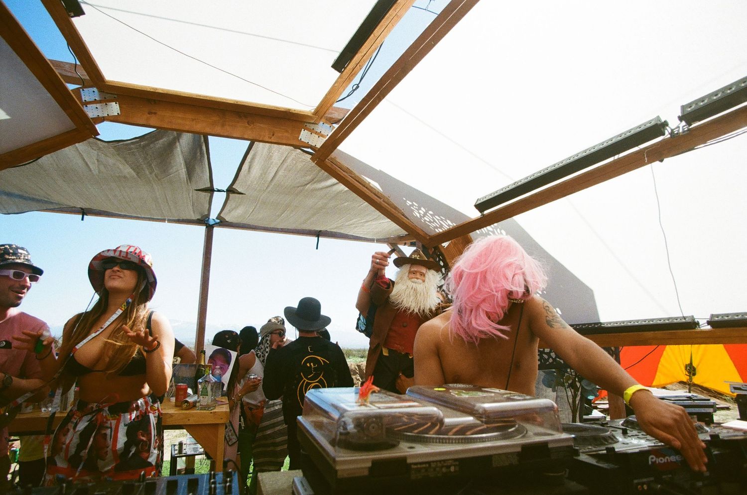 Pink-wigged DJ performing under a canopy at a sunny outdoor festival with costumed crowd.