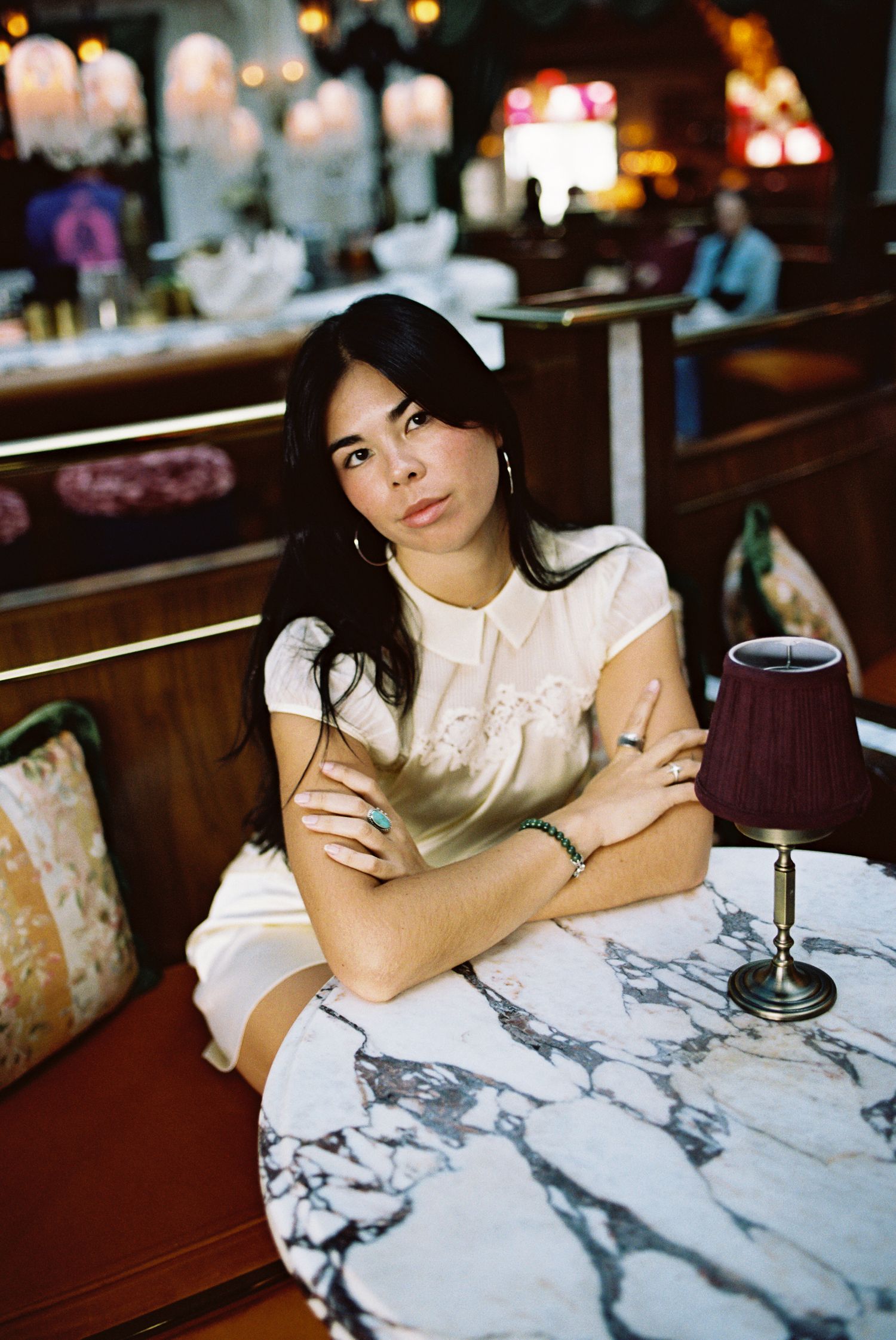Woman with dark hair in white dress sits arms crossed at marble table in elegant restaurant.