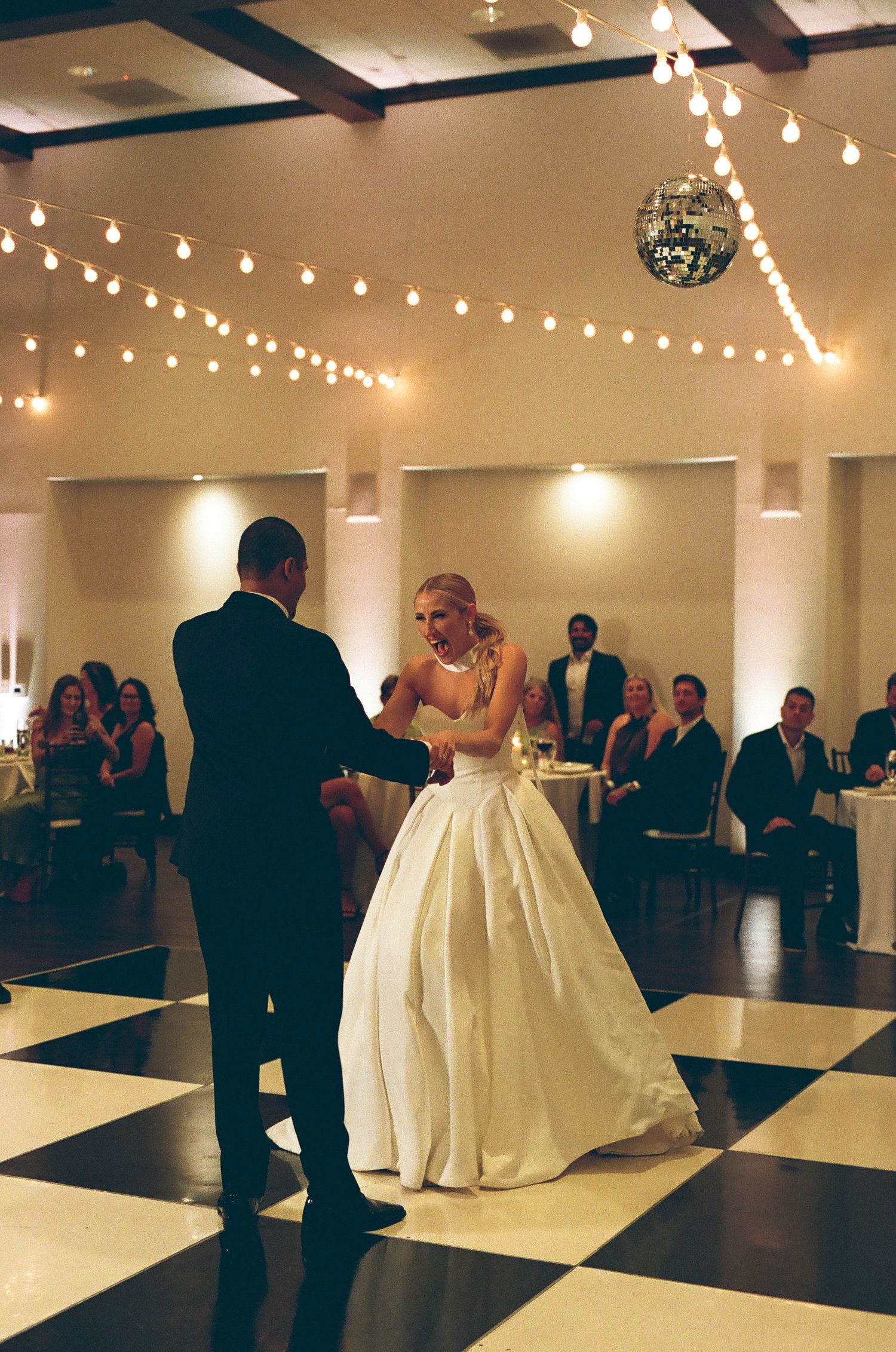 Bride and groom sharing first dance on black and white checkered floor under string lights and disco ball.