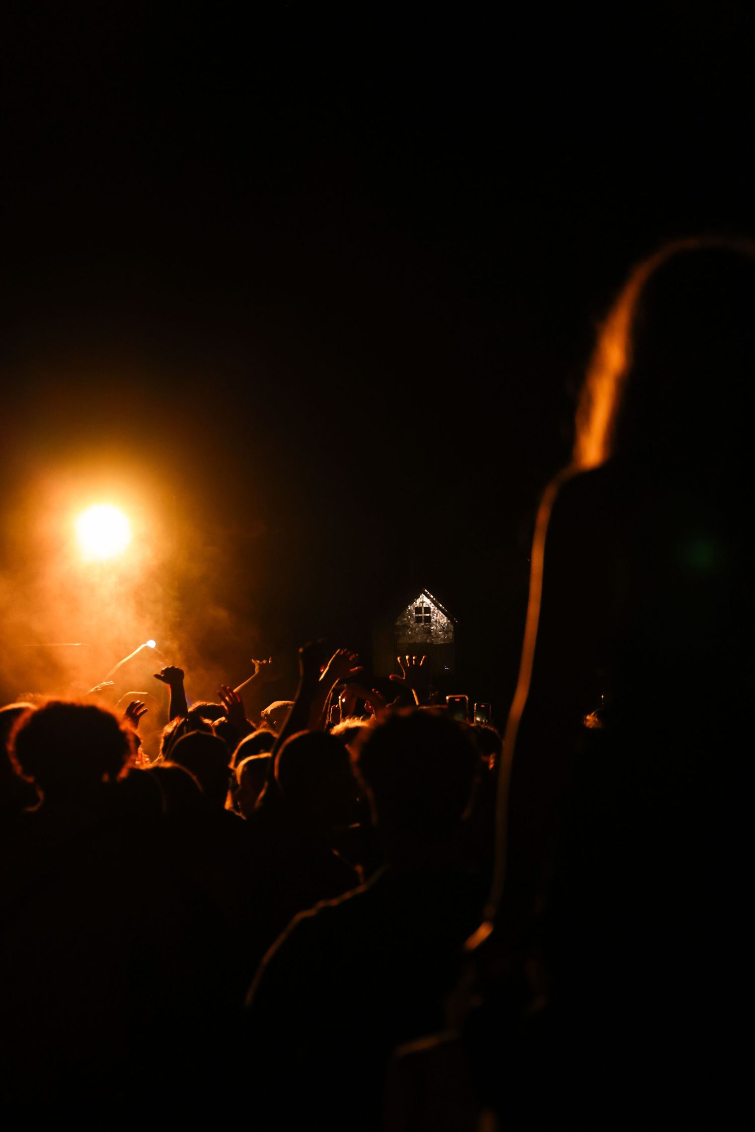 Concert crowd with raised hands illuminated by warm orange stage lights in the dark.