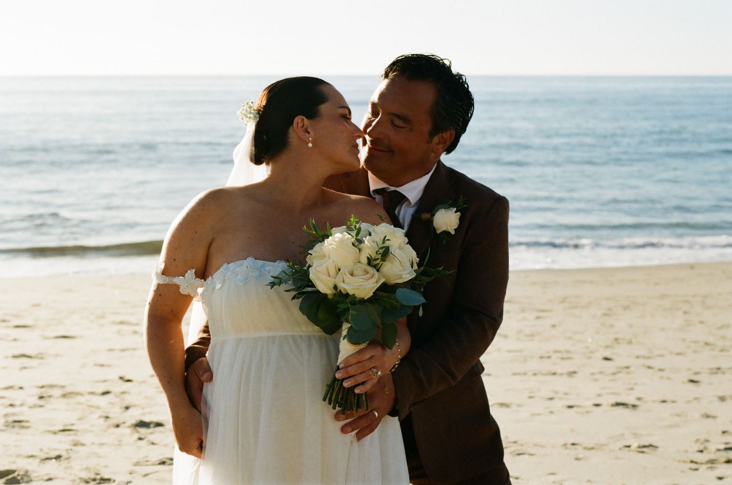 Bride and groom share a romantic kiss on a sandy beach, bride holding white bouquet.