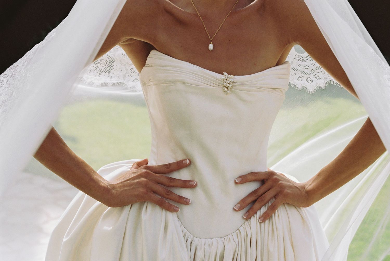 Bride in strapless ivory wedding gown with lace details, hands on hips, wearing delicate necklace.